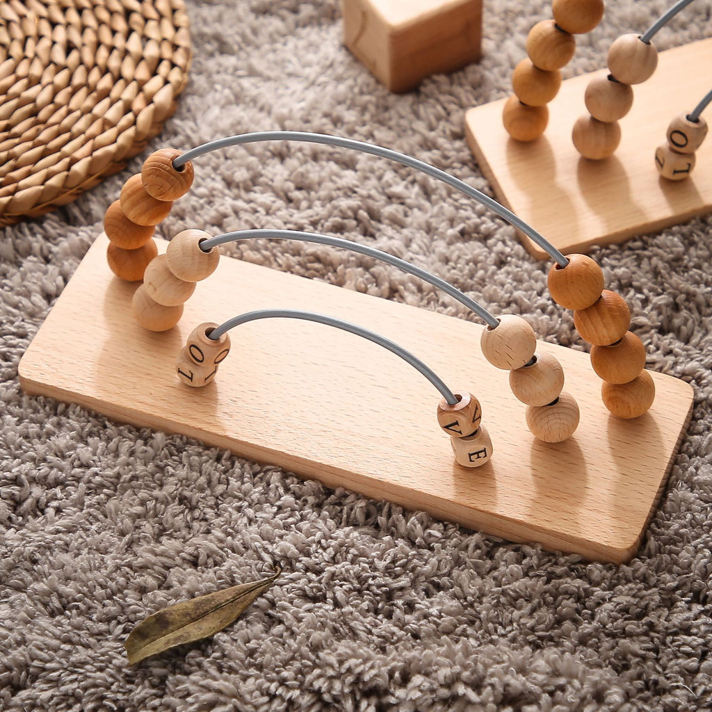 Wooden counting frame resting on a soft grey rug, showing natural beech beads threaded on curved wire arcs and small lettered blocks