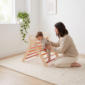 Parent kneeling beside the orange Pikler triangle spotting their toddler as they climb