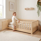 Toddler stepping into the twin size oak Montessori floor bed through the open front-left gate in a bright nursery